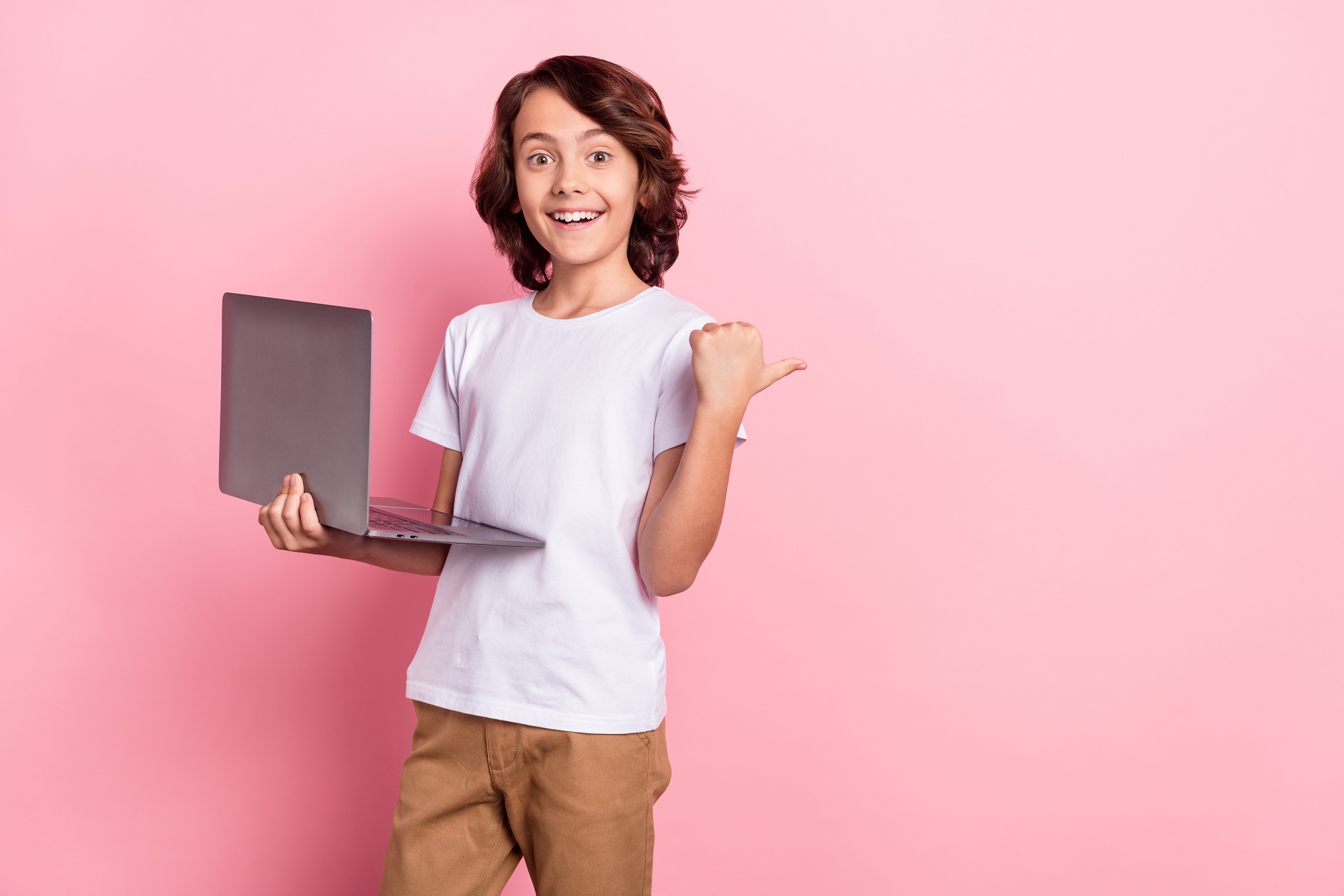Photo of small funny pupil boy point empty space hold laptop wear white t-shirt isolated on pastel pink color background.